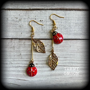May include: A pair of gold-colored earrings featuring a ladybug design with red and black enamel. Each earring has a dangling leaf charm and a ladybug charm. The earrings are on a white wooden surface. The text "TAMMY GMSON" is visible.