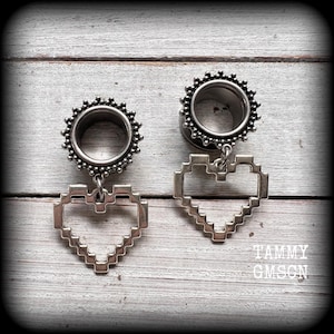 May include: A pair of silver-toned ear plugs featuring a circular tunnel design with a beaded outer edge. Each plug is adorned with a dangling, open-frame pixelated heart charm. The text TAMMY GMSCN is visible in the lower right corner of the image.