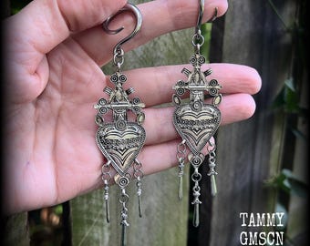 May include: Silver earrings with a heart-shaped design and dangling charms. The earrings have intricate detailing and a hook closure. The image also includes the text "TAMMY GMSCN".