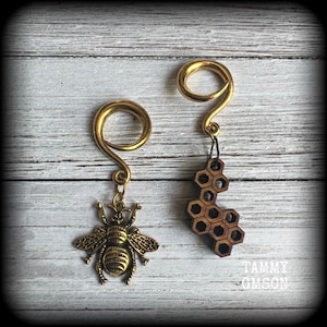 May include: Two gold-coloured spiral earrings. One has a detailed bee charm, the other a wooden honeycomb design. The earrings are shown against a light-coloured, textured background.