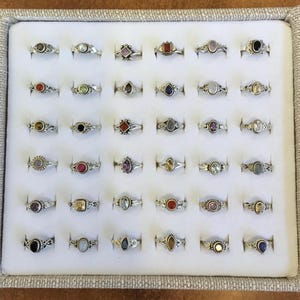 Può includere: Una collezione di trenta anelli in argento disposti in una griglia su un vassoio espositore bianco. Ogni anello presenta una pietra preziosa unica di varie forme, tra cui ovale, rotonda e a goccia, incastonata in fasce di metallo argentato.