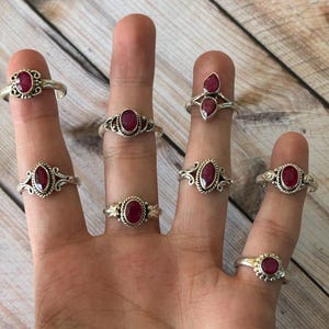 May include: A set of seven silver rings with red ruby gemstones. The rings are displayed on a person's fingers.