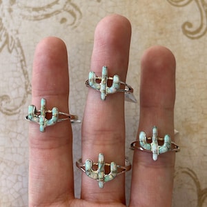 May include: Four silver rings with a white opal cactus design. The rings are stacked on a hand.