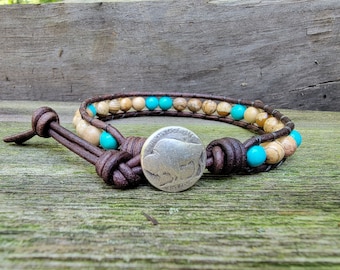 May include: A brown leather bracelet with a silver buffalo coin charm. The bracelet is adorned with light brown and turquoise beads.