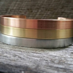 May include: A set of three stacked metal bracelets. The top bracelet is copper, the middle bracelet is gold, and the bottom bracelet is silver.