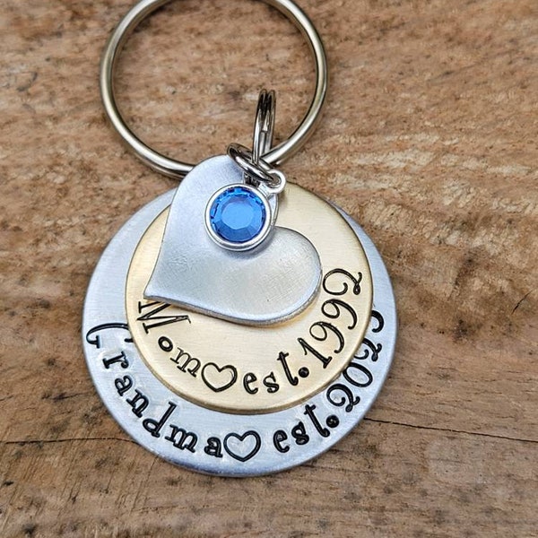 May include: Silver and gold tone keychain with a heart charm and a blue rhinestone. The keychain has the text "Mom est. 1999" and "Grandma est. 2023" engraved on it.