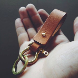 Puede incluir: Un llavero de cuero marrón con un anillo en D de latón y un remache de latón. El llavero está sostenido en una mano.