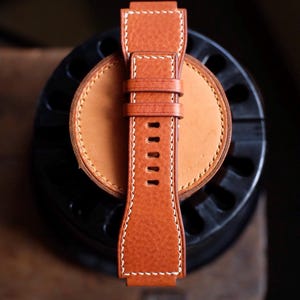 May include: Close-up of a brown leather watch strap with white stitching. The strap has a textured surface and a buckle. The watch strap is resting on a light brown leather circle.