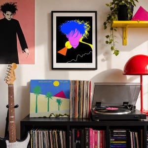 May include: A framed pop art print featuring a stylized face with blue hair, a pink face, and yellow accents. The print hangs above a record player and a vinyl record album cover with palm trees and the text "Boys Don't Cry".