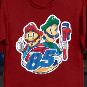 May include: Maroon t-shirt featuring a graphic of two cartoon characters, one in a red hat and the other in a green hat, with the number 85. The red character gives a thumbs up. The green character holds a wrench. Text at the bottom of the shirt reads "COMES IN BOTH MALE AND FEMALE SIZES ARTIST MOCK UP ACTUAL SIZE AND COLORS OF SHIRT AND DESIGN MAY VARY."