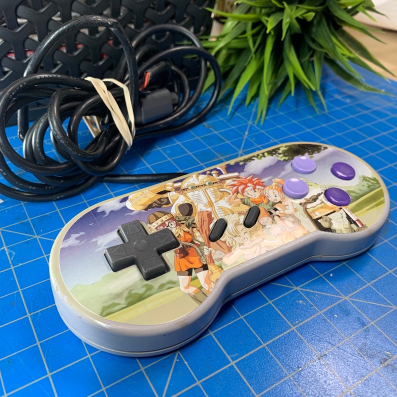 May include: A gray Super Nintendo controller with a colorful graphic featuring characters from a video game. The controller has a directional pad, black buttons, and purple buttons. A black cable is attached.