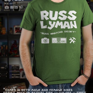 May include: Green t-shirt with the text "RUSS LYMAN" in white block letters. Below the name is "RUSS WHATCHA DOIN'?" and icons of a camera, game controller, and wrench. The shirt is a crew neck style.