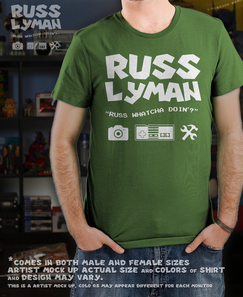 May include: Green t-shirt with the text "RUSS LYMAN" in white block letters. Below the name is "RUSS WHATCHA DOIN'?" and icons of a camera, game controller, and wrench. The shirt is a crew neck style.