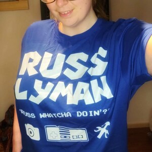May include: A blue t-shirt with the text "RUSS LYMAN" in large white letters. Below the name is the phrase "RUSS WHATCHA DOIN'?" and graphic icons of a camera, a game controller, and a wrench.