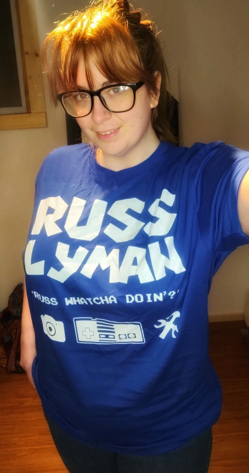 May include: A blue t-shirt with the text "RUSS LYMAN" in large white letters. Below the name is the phrase "RUSS WHATCHA DOIN'?" and graphic icons of a camera, a game controller, and a wrench.