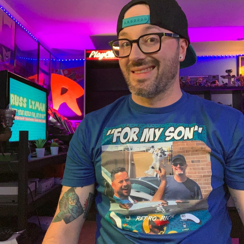 May include: Blue t-shirt with the phrase "FOR MY SON" above a photo of two men. The shirt has a Retro Rick logo. The person wearing the shirt is also wearing glasses and a black baseball cap.