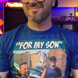 May include: Blue t-shirt with the text "FOR MY SON" in white. The shirt features a graphic of two men in a car and the words "Retro Rick". The person in the image is wearing a black cap and glasses.