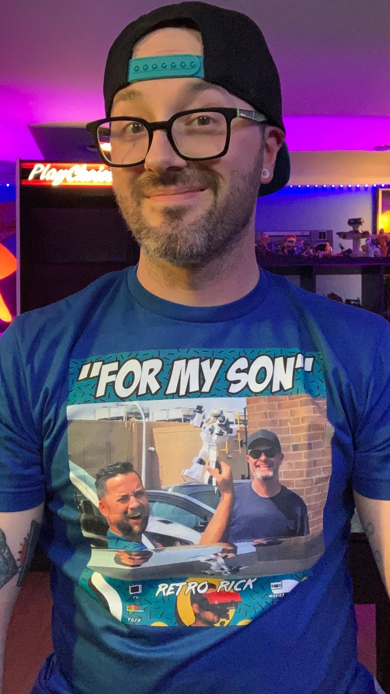 May include: Blue t-shirt with the text "FOR MY SON" in white. The shirt features a graphic of two men in a car and the words "Retro Rick". The person in the image is wearing a black cap and glasses.