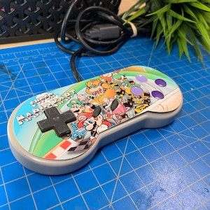 May include: A gray Super Nintendo controller featuring a colorful Super Mario Kart design. The controller has a black directional pad, purple buttons, and a cable attached. The design includes various characters from the game.