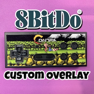 May include: A rectangular 8BitDo game controller featuring a custom Contra video game graphic overlay. The controller has a black D-pad, a central horizontal button, and four circular black action buttons set against a pixelated jungle scene.