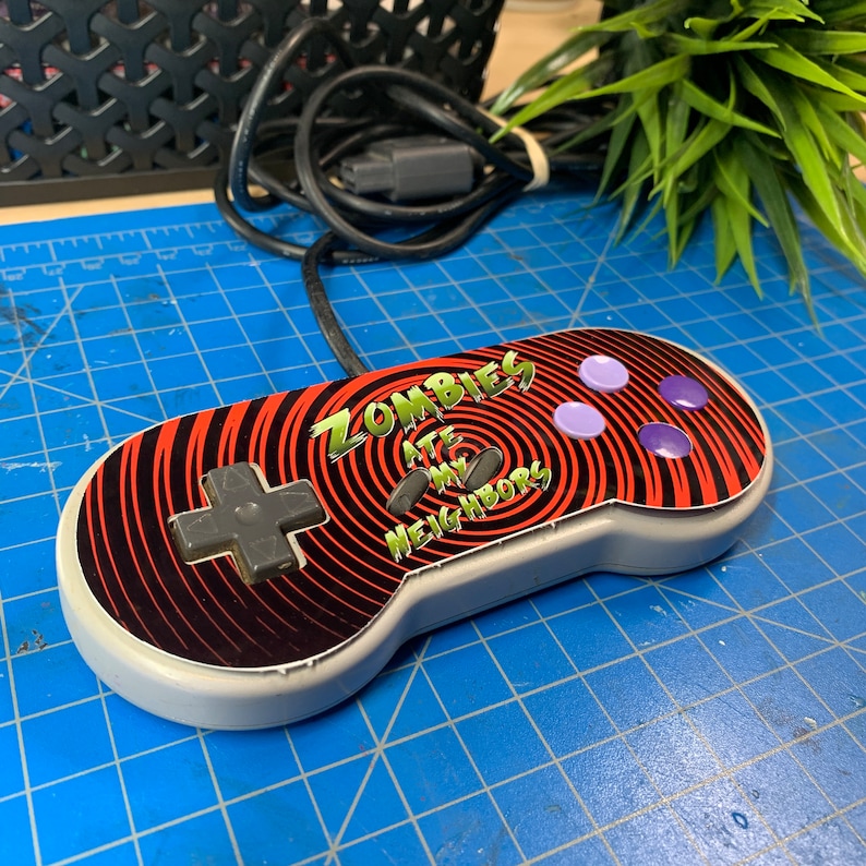 May include: A retro video game controller with the text "Zombies Ate My Neighbors" on the face. The controller has a gray body, a black directional pad, and purple and lavender buttons. The face has a red and black spiral design.