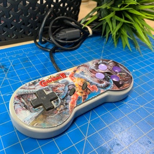 May include: A gray Super Nintendo controller with a colorful graphic from the game Castlevania III. The controller has a black directional pad, two black buttons, and four purple buttons. A black cable extends from the controller.