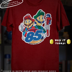 May include: Maroon t-shirt featuring a graphic of two cartoon plumbers with the number 85. One plumber gives a thumbs up. The text "BUY IT TODAY!" is displayed. The shirt has the text "STAY UP TO DATE ON NEW ITEMS FROM THIS SHOP!"