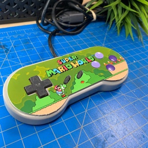 May include: A gray Super Nintendo controller with a green and white Super Mario World design. The controller features a black directional pad, purple buttons, and the game's logo. A black cable extends from the top of the controller.