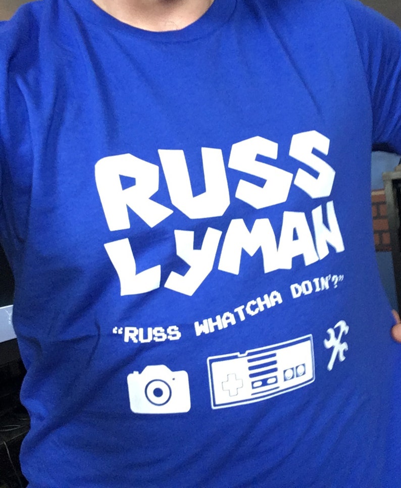 May include: A royal blue t-shirt with the name "RUSS LYMAN" in large white block letters. Below the name, the text reads "RUSS WHATCHA DOIN?" with a camera, game controller, and wrench icons.
