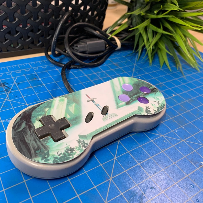 May include: A gray Super Nintendo controller with a custom design featuring a forest scene and a sword. The controller has a directional pad, four face buttons, and purple buttons. The controller is on a blue cutting mat.
