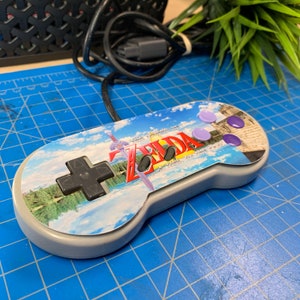 May include: A gray Super Nintendo controller with a custom design featuring the text "ZELDA" and a landscape scene. The controller has purple buttons and a black directional pad. A black cable is attached.
