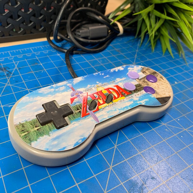 May include: A gray Super Nintendo controller with a custom design featuring the text "ZELDA" and a landscape scene. The controller has purple buttons and a black directional pad. A black cable is attached.