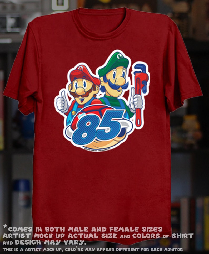 May include: Maroon t-shirt featuring a graphic of two cartoon characters, one in a red hat and the other in a green hat, with the number 85. The red character gives a thumbs up. The green character holds a wrench. Text at the bottom of the shirt reads "COMES IN BOTH MALE AND FEMALE SIZES ARTIST MOCK UP ACTUAL SIZE AND COLORS OF SHIRT AND DESIGN MAY VARY."
