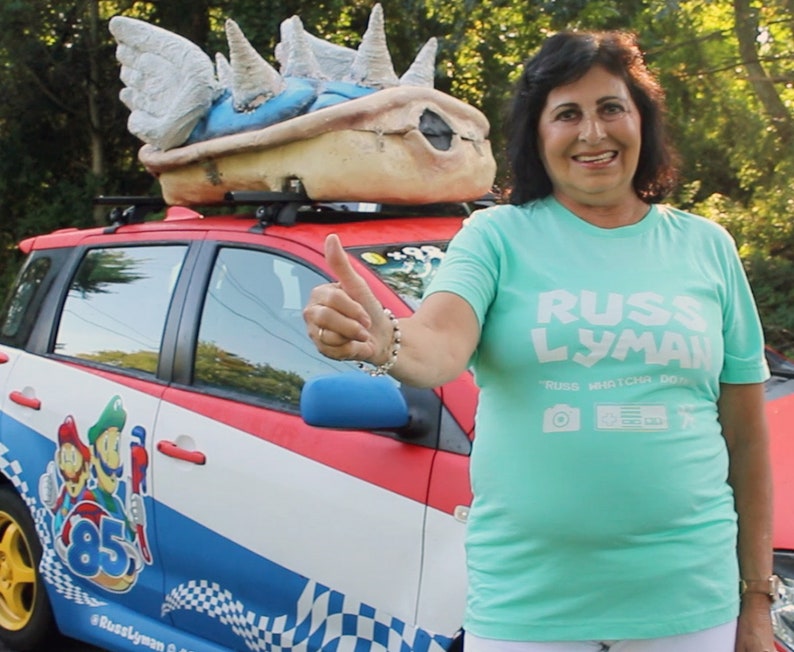 May include: A red, white, and blue car with a large Bowser shell on top. The car features Mario and Luigi graphics and the number 85. The person is wearing a light green t-shirt with the text "RUSS LYMAN".