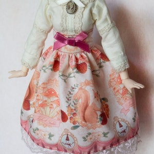 Può includere: Una bambola che indossa una blusa bianca con pizzo e un fiocco rosa, una gonna con stampa di scoiattolo e funghi e un bordo di pizzo bianco. La gonna ha un nastro rosa intorno alla vita.