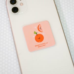 Könnte beinhalten: Ein quadratischer Aufkleber mit hellrosa Hintergrund auf der Rückseite eines weißen Smartphones. Der Aufkleber zeigt eine Cartoon-Orange mit lächelndem Gesicht, eine Orangenscheibe und den Text "YOU'RE CITRUS-LY AMAZING!" in brauner Schrift.