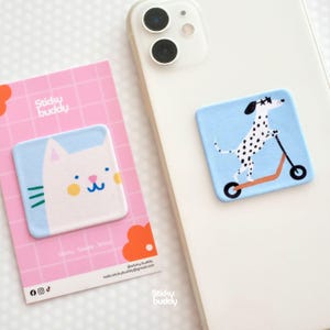 Op de afbeelding: Twee telefoonaccessoires. Eén heeft een witte kattenkop met blauwe accenten op een lichtblauwe achtergrond, de andere een Dalmatische hond op een scooter op een lichtblauwe achtergrond. Beide bevinden zich op een witte telefoon en een roze rasterkaart.