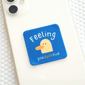 Op de afbeelding: Een vierkante blauwe sticker bevestigd op een witte smartphone. De sticker bevat een afbeelding van een gele eendenkop met een oranje snavel en de witte tekst "Feeling" erboven en "producktive" eronder, waarbij de "u" in "producktive" oranje is.