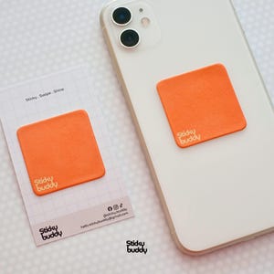 Puede incluir: Un Sticky Buddy cuadrado naranja adherido a un teléfono inteligente blanco. Un Sticky Buddy a juego está en una tarjeta blanca pequeña con el texto "Sticky. Swipe. Shine." y el logotipo de Sticky Buddy. El fondo es un patrón de cuadrícula blanca.
