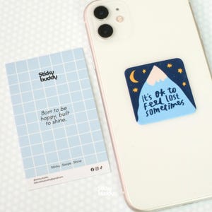 Może przedstawiać: Biały smartfon z kwadratową naklejką przedstawiającą górę, księżyc i gwiazdy oraz napis It's OK to feel lost sometimes. Obok znajduje się jasnoniebieska karta w kratkę z napisem Sticky Buddy, Born to be happy, built to shine.