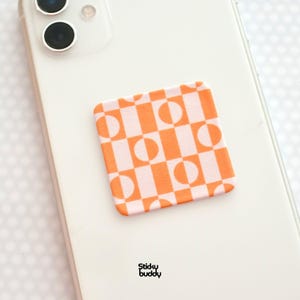 Op de afbeelding: Een vierkante telefoonhouder met een oranje en wit geometrisch patroon van halve cirkels en vierkanten. Het telefoonaccessoire is bevestigd aan de achterkant van een witte smartphone. De woorden "Sticky buddy" staan onderaan gedrukt.