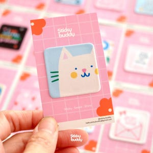 Op de afbeelding: Een vierkante sticker met een illustratie van een witte kat met blauwe ogen, een roze neus en gele wangen op een lichtblauwe achtergrond. De sticker zit op een roze kaart met een wit ruitpatroon en de tekst Sticky buddy.