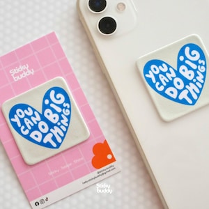 Op de afbeelding: Een vierkante, gebroken witte zelfklevende telefoonhouder met een blauw hartmotief en de witte tekst "YOU CAN DO BIG THINGS". De houder wordt getoond op een roze kaart met rasterpatroon en is bevestigd op de achterkant van een witte smartphone.