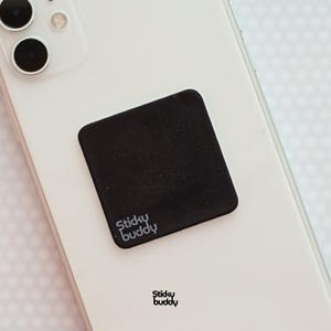 Op de afbeelding: Een vierkante zwarte zelfklevende pad met afgeronde hoeken bevestigd aan de achterkant van een witte smartphone. De pad is voorzien van de witte tekst Sticky Buddy in de linkerbenedenhoek. Een klein logo met de tekst Sticky Buddy is onder de telefoon.