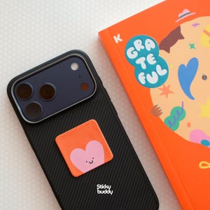 Op de afbeelding: Een zwarte smartphonehoes met een diagonaal textuurpatroon en een vierkante oranje zelfklevende grip met een roze hartfiguurtje met een lachend gezicht. De tekst Sticky buddy is in het wit onderaan de telefoonhoes gedrukt.