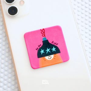 Op de afbeelding: Een vierkante roze telefoonhouder met een donkerblauwe lampafbeelding met drie lichtblauwe sterren, een lachend gezicht en de tekst Be the light. Het accessoire is bevestigd aan de achterkant van een witte smartphone met dubbele cameralens.