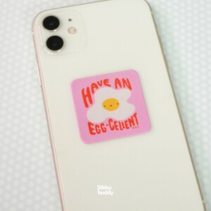 Op de afbeelding: Een witte smartphone met een vierkante roze sticker met een cartoon gebakken ei met een lachend gezicht. De tekst op de sticker luidt HAVE AN EGG-CELLENT DAY in rode letters. De sticker is gecentreerd op de achterkant van de telefoon geplaatst.