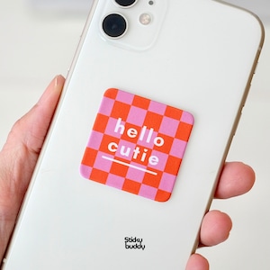Może przedstawiać: Kwadratowa naklejka w różowo-pomarańczową kratkę przyklejona do tyłu białego smartfona. Naklejka zawiera biały napis "hello CUTIE" z małym serduszkiem nad literą i oraz poziomą linią znajdującą się bezpośrednio pod tekstem.