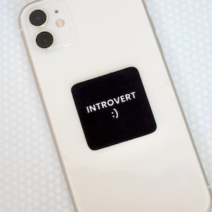 Op de afbeelding: Een witte smartphone met een vierkante zwarte sticker in het midden van de achterkant. De sticker bevat de witte tekst "INTROVERT" in hoofdletters, gevolgd door een smiley-emoticon bestaande uit een dubbele punt en een haakje.