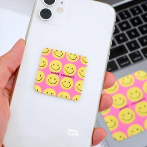 Op de afbeelding: Een witte smartphone met een vierkante sticker met een roze achtergrond en een herhalend patroon van gele smileygezichten. De tekst HAPPY THOUGHTS is in het zwart op de sticker gedrukt. Een bijpassende sticker is zichtbaar op de laptop.